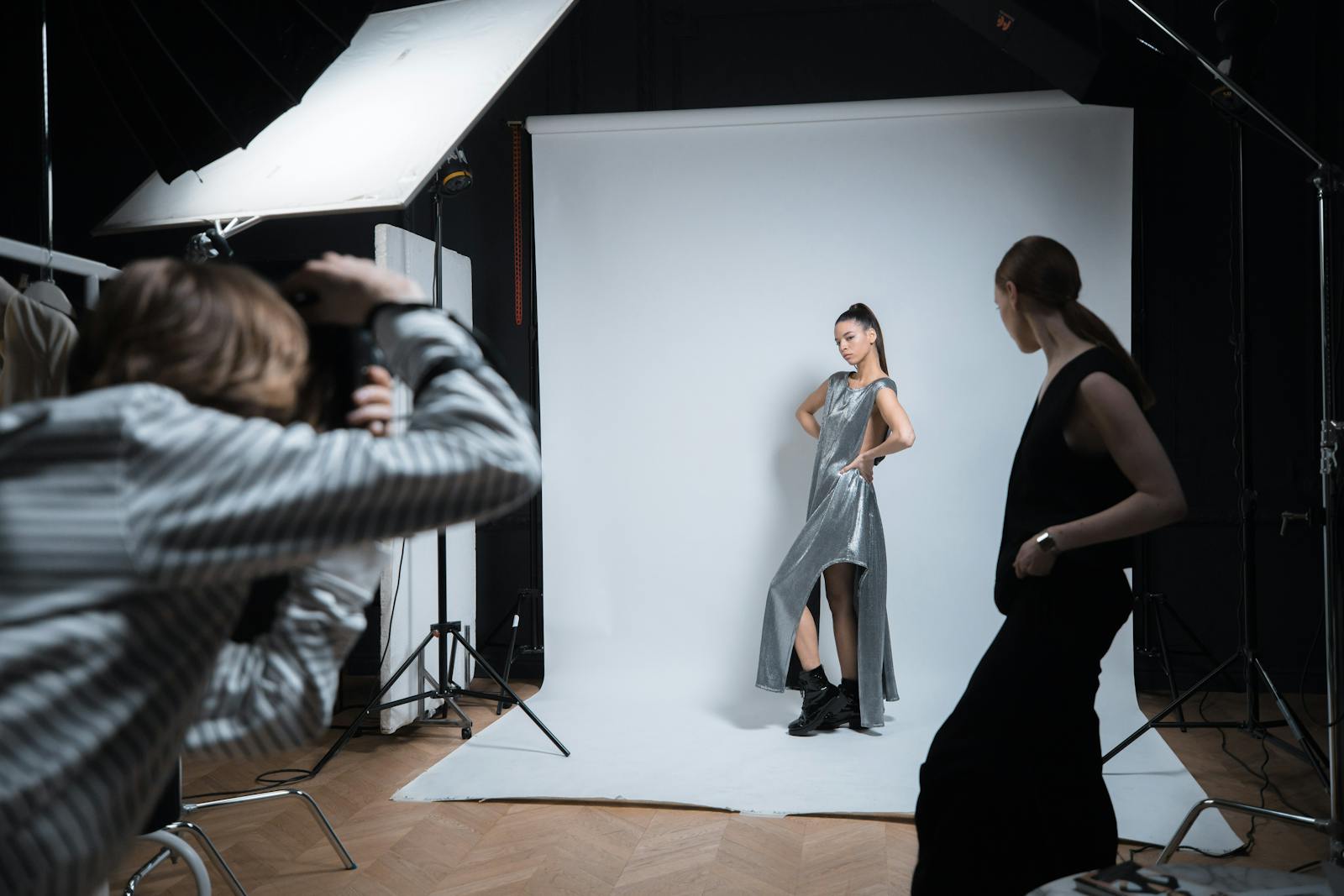A working set in the Brivon Brooklyn studio — two stylists pinning a garment on a model while a photographer reviews tethered captures on a calibrated monitor.