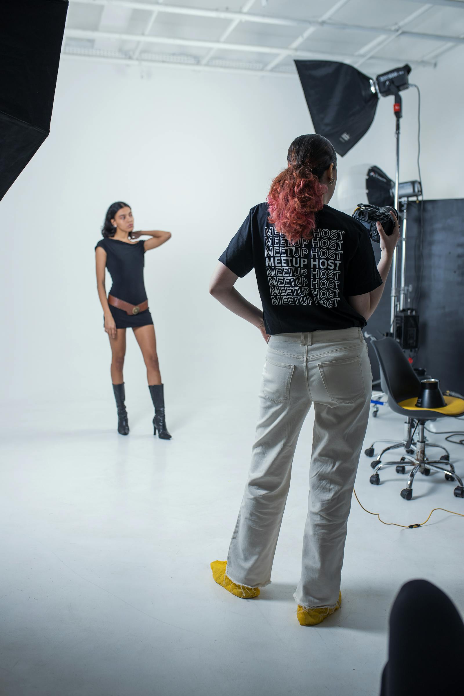 A daylight photography studio with high ceilings, a single grey seamless rolled down, and a stylist arranging garments on a rack against the far wall.