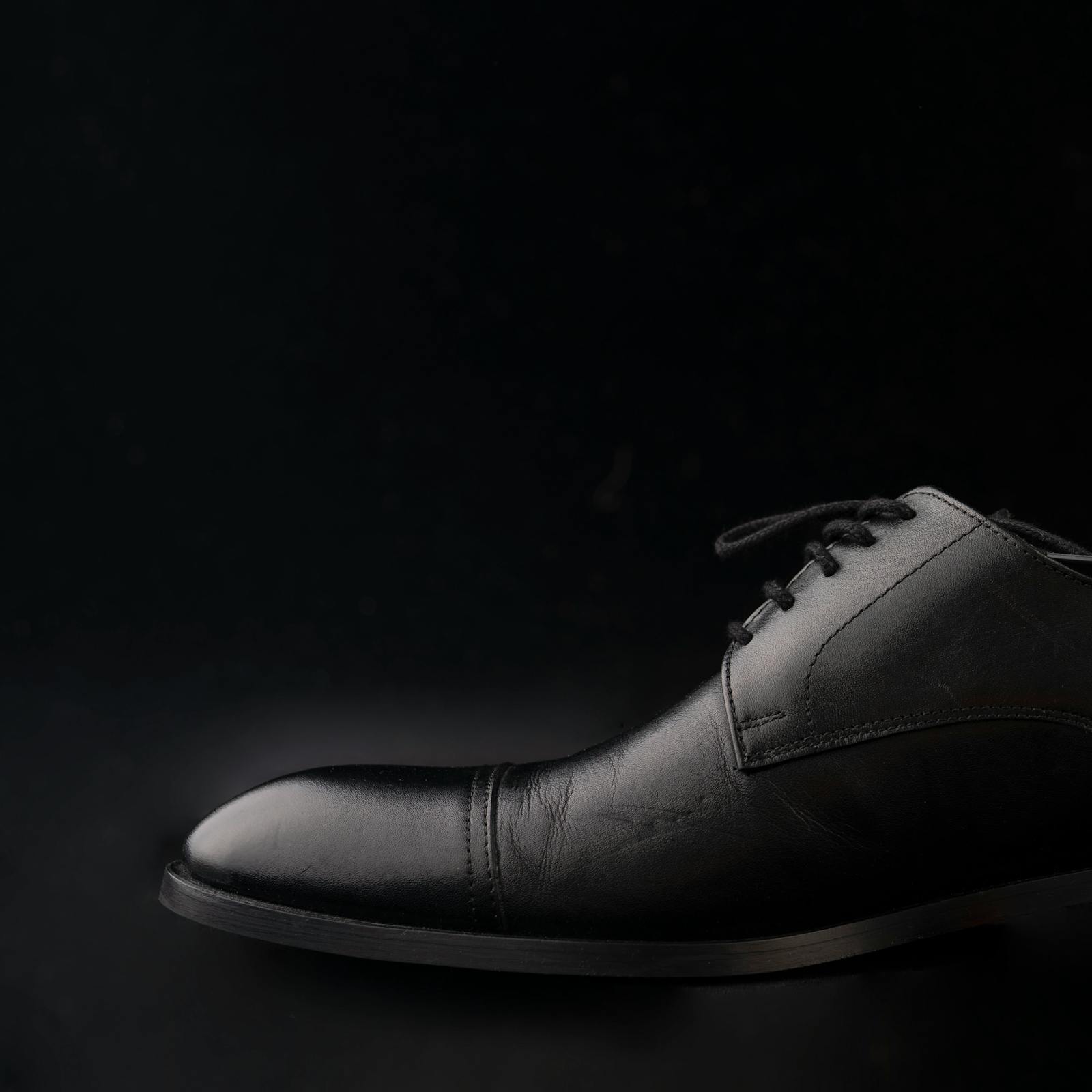 A pair of leather oxfords on a marble plinth, lit by a single hard light to throw a sharp shadow against a charcoal backdrop.